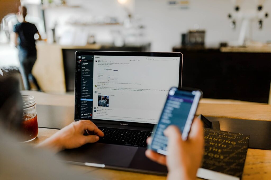 A person sits at a coffee shop, using a laptop while holding a smartphone, immersed in their work.