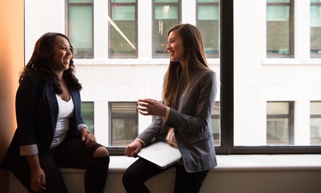 Two women engaged in conversation by an office window, sharing ideas and collaborating in a professional setting.