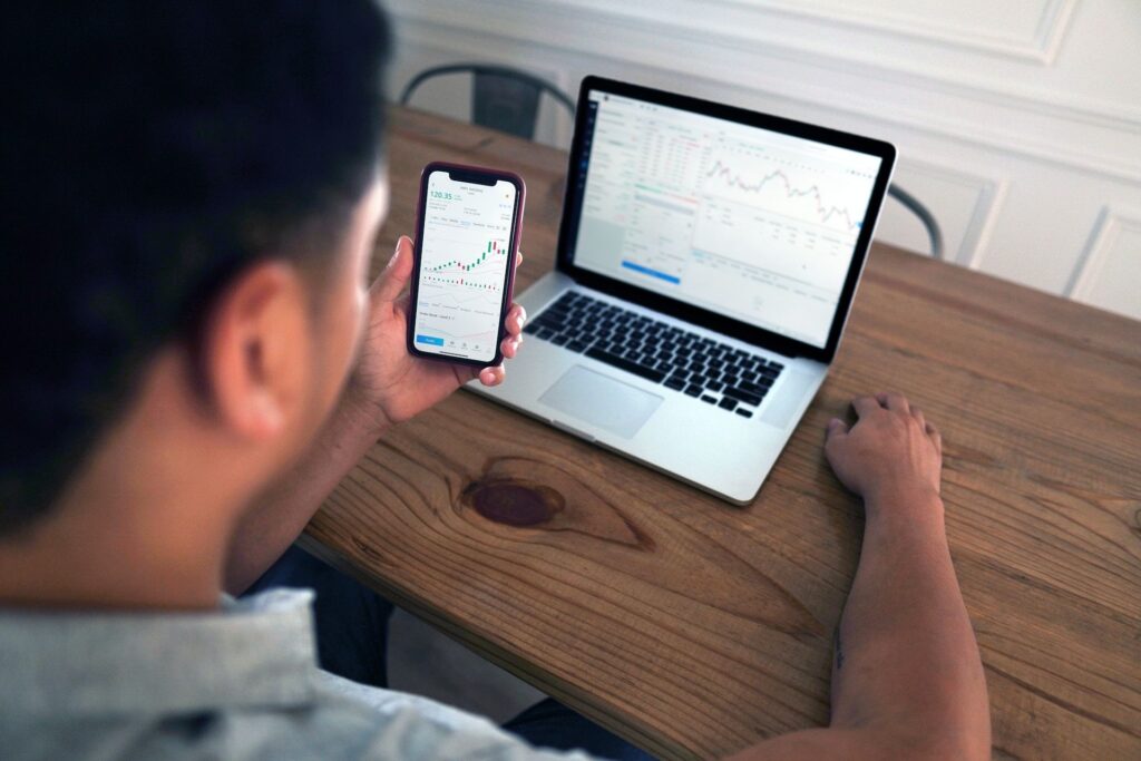 A man monitors the stock market using a smartphone and laptop, focused on financial data and market trends.