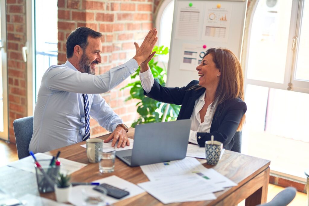 Two business professionals celebrate success with a high five at a desk, showcasing teamwork and achievement in the workplace.