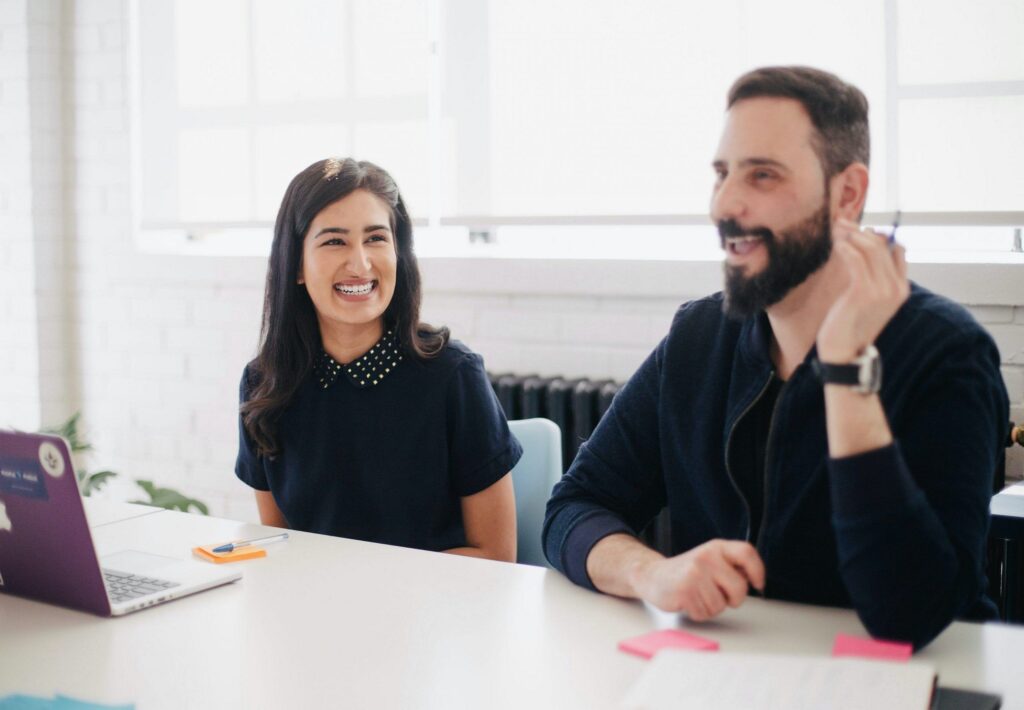 A man and woman are seated at a table, engaged with a laptop in front of them, discussing or collaborating on a project.